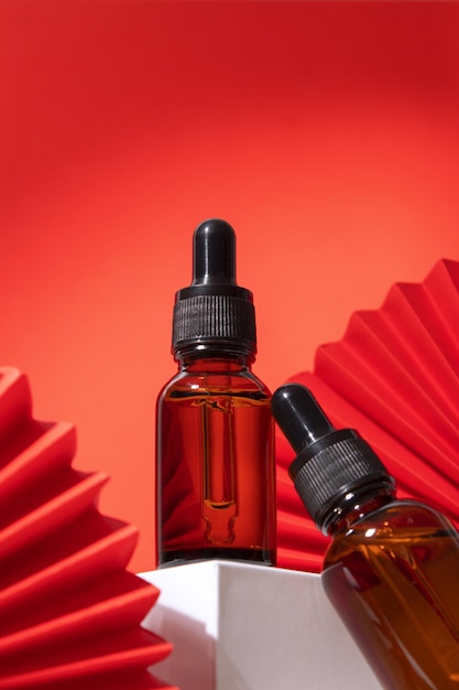 Beauty serum in a dropper bottle on a podium with geometric shapes paper fan on a red background