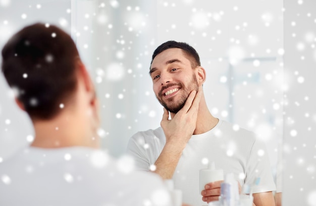 아름다움, 면도, 몸단장, 겨울, 사람 개념 - 거울을 보고 웃고 있는 젊은 남자가 눈 위에 집 화장실에서 애프터셰이브를 바르고 있습니다.