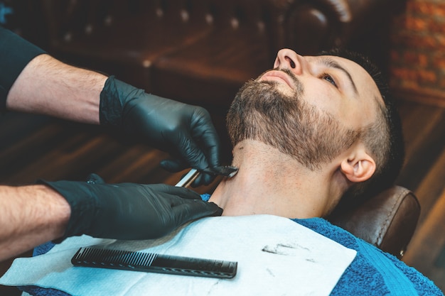 Beauty shop for men. Shaving a beard in a barbershop. Barber cuts his beard with a razor and clipper. close up Brutal haircuts. Hairdresser equipment. Selective focus.