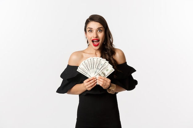 Beauty and shopping concept. Excited woman in black dress, showing money prize and staring happy at camera, standing over white background