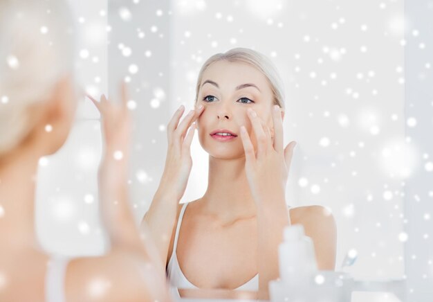 写真 美容皮膚ケア人間のコンセプト - 笑顔の若い女性が顔にクリームを塗り自宅の浴室で雪の上に鏡を見ています