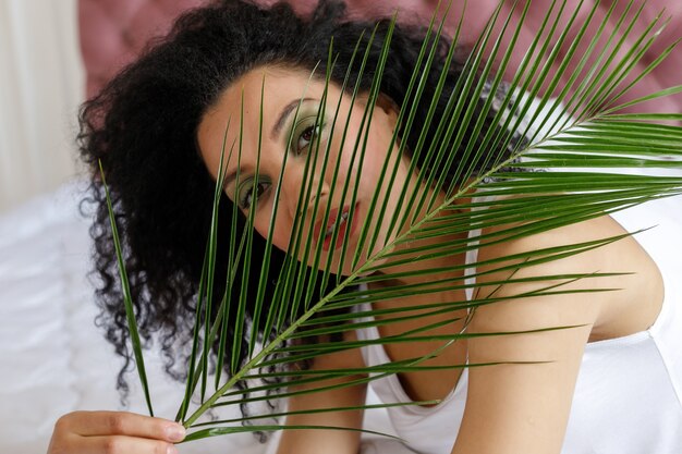 Photo beauty, skin care and people concept - close up portrait of beautiful mixed race woman. portrait of an african woman, lying on a snow-white bed looking at the camera. girl and palm branch.