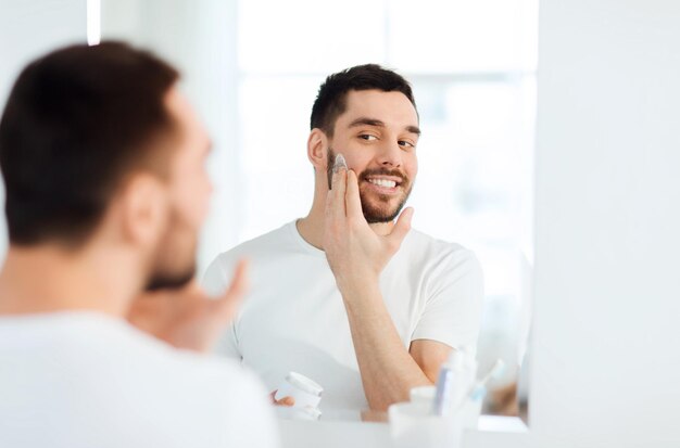 красота, уход за кожей и концепция людей - улыбающийся молодой человек наносит крем на лицо и смотрит в зеркало в домашней ванной