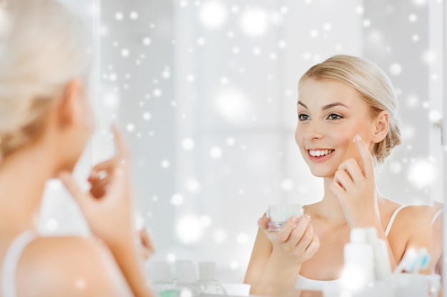 beauty, skin care and people concept - smiling young woman applying cream to face and looking to mirror at home bathroom over snow