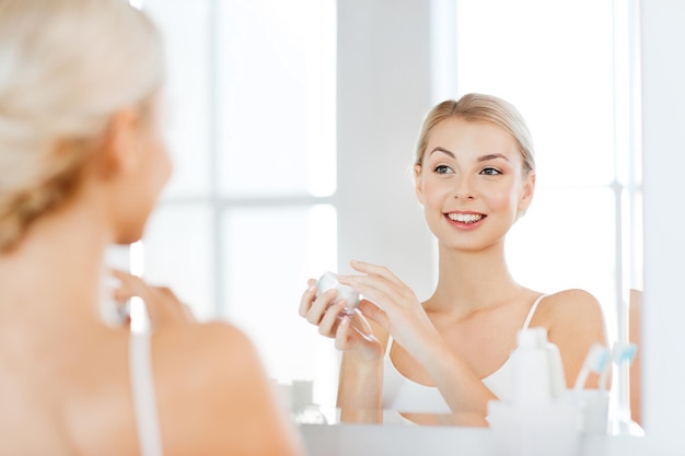 Photo beauty, skin care and people concept - smiling young woman applying cream to face and looking to mirror at home bathroom