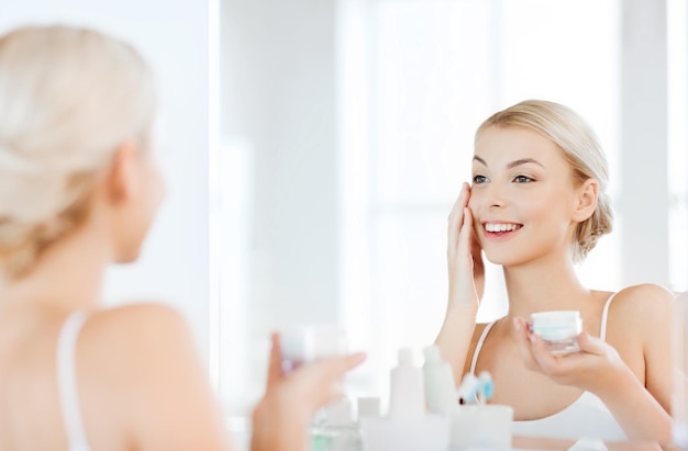 Красота, уход за кожей и люди концепции - улыбается молодая женщина, применяя крем для лица и глядя в зеркало в ванной комнате дома