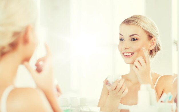 beauty, skin care and people concept - smiling young woman applying cream to face and looking to mirror at home bathroom