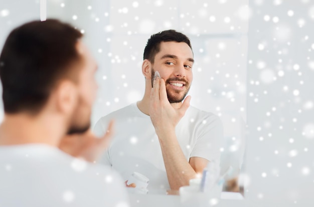 beauty, skin care, winter and people concept - smiling young man applying cream to face and looking to mirror at home bathroom over snow