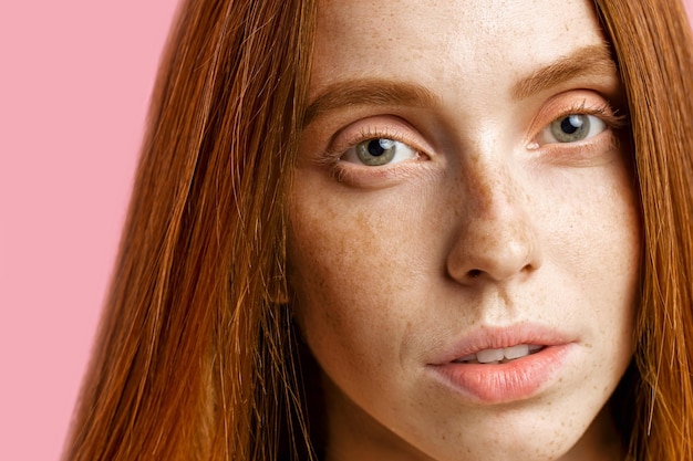 Beauty, skin and hair care concept. Close up shot of beautiful redhead girl with perfect clean skin, with freckles, no make up, looking at camera, standing over pink background.