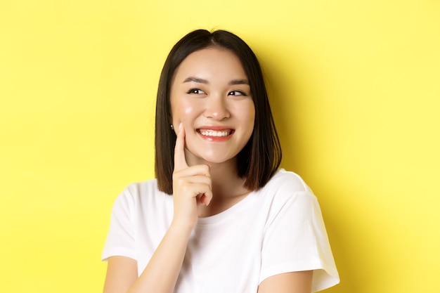 Bellezza e cura della pelle. primo piano di una giovane donna asiatica con capelli scuri corti, pelle sana e luminosa, fossette sorridenti e toccanti sulle guance, in piedi su sfondo giallo.