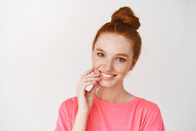 Beauty and skincare Closeup of cute teenage girl with ginger hair combed in bun smiling with white teeth showing glowing skin no makeup on face standing over white background