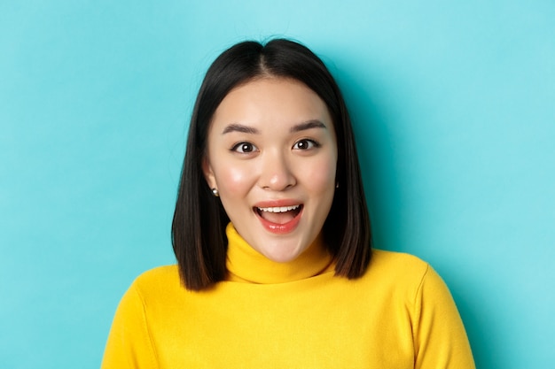 Beauty and skincare concept. Close up of surprised asian female raising eyebrows and look amused at camera, look at something interesting, standing over blue background.