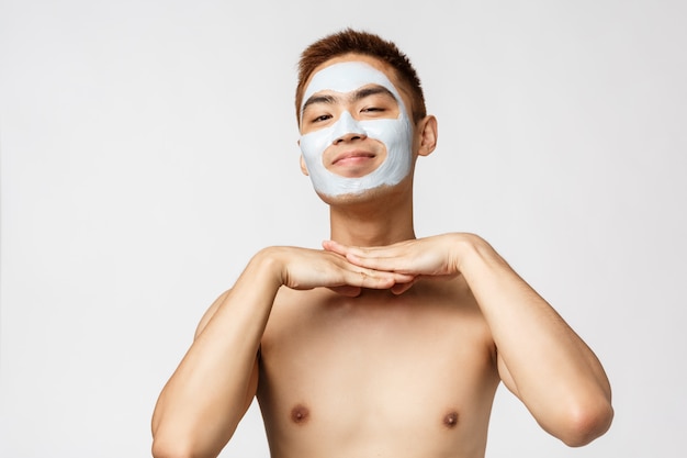 美容、スキンケア、スパのコンセプトです。素敵で愚かな裸のアジア人男性の肖像画、満足して笑顔、顔のマスククリームを着て、肌の世話をする化粧品、立っている白い壁