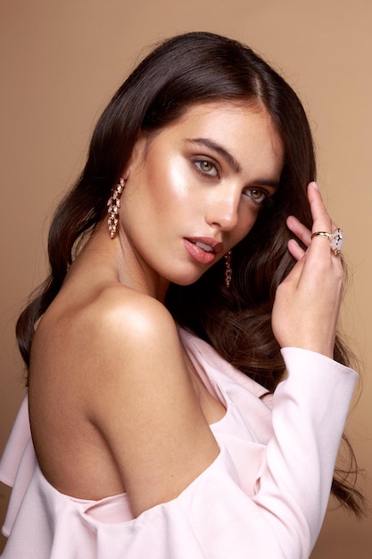 裸の肩、茶色の髪、茶色の背景の上にピンクの石の指輪とイヤリングを身に着けている女性の美容スタジオの肖像画。