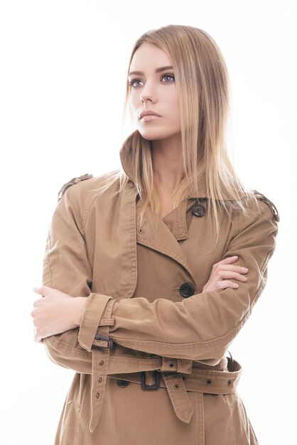 Beauty in style. Attractive young woman in coat keeping arms crossed and looking away while standing against white background