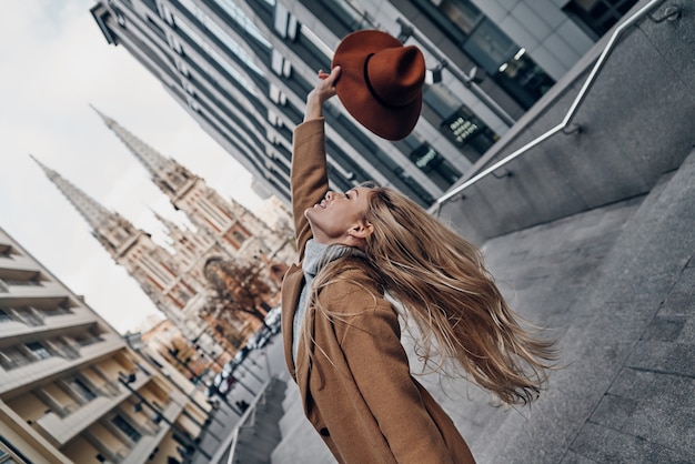 Beauty and style. Attractive young woman holding hat and smiling while spending carefree time in the city