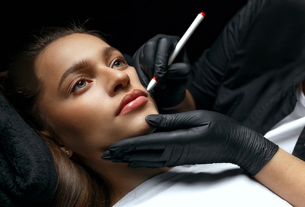 Beauty stylist making lips shape before permanent makeup procedure at the cosmetic salon