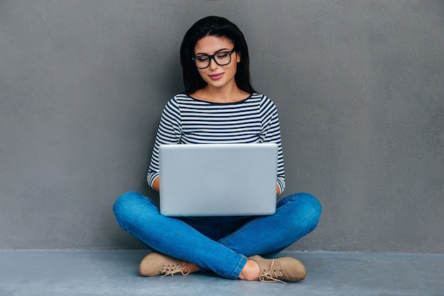 Beauty surfing web. Attractive young woman carrying laptop on her knees and looking at it while sitting on the floor and against grey background