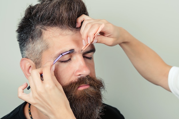 Beauty treatment. Over the shoulder view of handsome male plucking eyebrows while in beauty salon. Female hands plucking eyebrow tweezers to relaxed bearded brutal man isolated over gray background