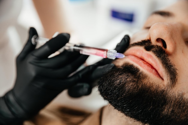 Косметические процедуры для стильного молодого человека с бородой Косметолог делает инъекции красоты в лицо мужчины