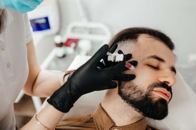 Beauty treatments for a stylish young man with a beard Beautician makes beauty injections in a man's face