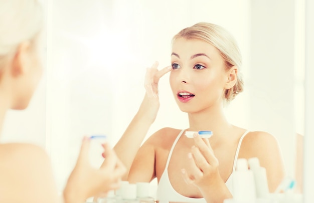 beauty, vision, eyesight, ophthalmology and people concept - young woman putting on contact lenses at mirror in home bathroom