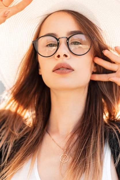 Beauty vogue portrait pretty young gorgeous woman with sexy lips in youth glasses in black jacket in elegant hat on bright sunny day. Stylish girl fashion model enjoys in  warm rays of summer sun.