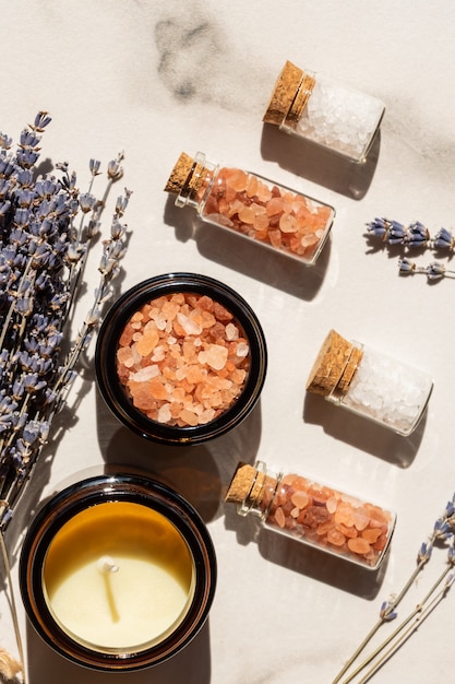 Beauty and wellness concept - sea salt in glass bottles and candle on wooden tray and bunch of lavender on marble background