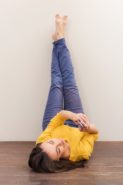 Beauty with mobile phone. Beautiful young woman holding mobile phone while lying on the floor and leaning her legs at the wall