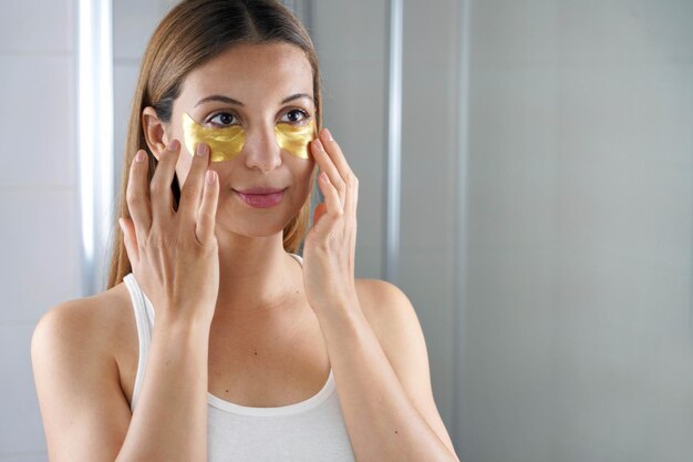 Photo beauty woman applying golden antiaging undereye mask looking herself in the mirror in the bathroom skin care girl touch patches of fabric mask under eyes to reduce eye bags