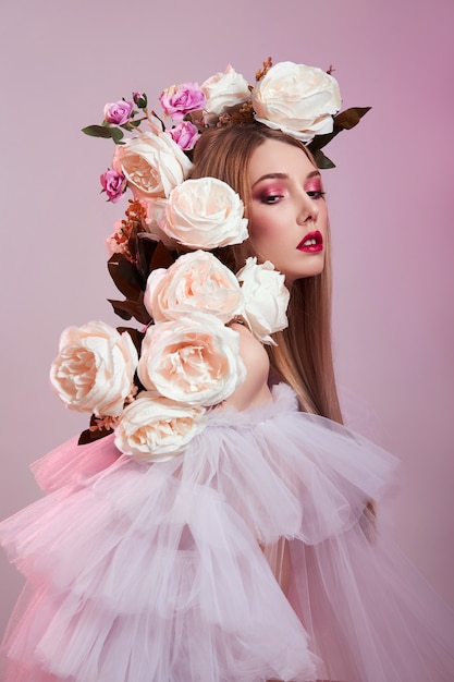 Beauty Woman bright red makeup, rose flowers on head woman. Art Girl in a transparent light dress on a pink wall. Professional bright makeup