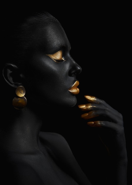 Beauty woman painted in black skin color body art, gold makeup lips eyelids, fingertips nails in gold color paint. Professional gold makeup