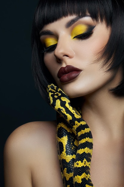 Beauty woman short haircut python yellow snake on her neck. a
yellow snake on the shoulders of a girl. beauty yellow eye shadow
makeup, dark burgundy lipstick