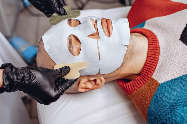 A beauty worker uses quartz gouache scrapers and massages a male in a facial skincare fabric mask with them