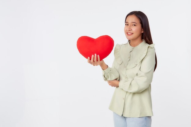 Bellezza giovane bruna asiatica donna che indossa la camicia verde in piedi tenere cuscino a forma di cuore rosso nella superficie dello studio isolato grigio chiaro