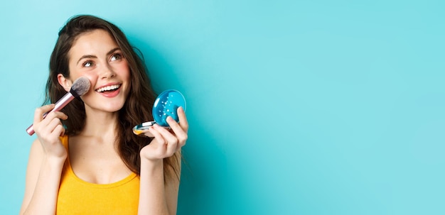 Beauty Young glamour girl applying make up with pocket mirror and brush smiling and gazing up at logo standing against blue background