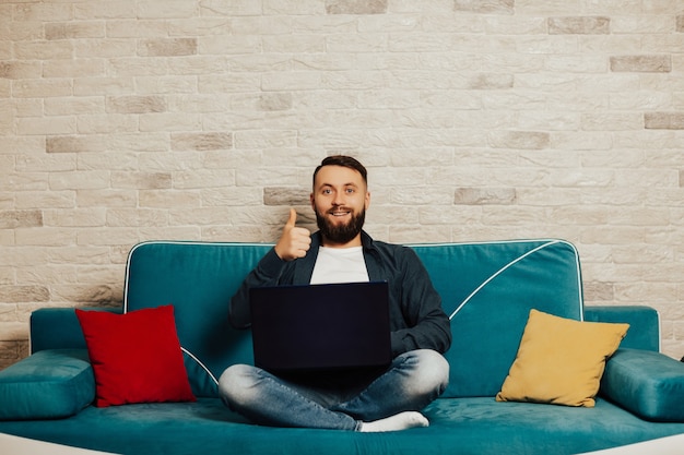 Bebaarde aantrekkelijke glimlachende man gebruikt op zijn laptop, thuis op de gezellige bank thuis zitten