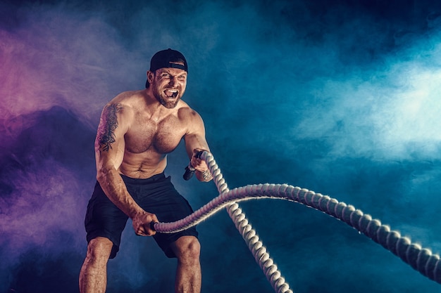 Bebaarde atletisch uitziende bodybulder trainen met gevechtstouw op donker met rook. Kracht en motivatie.