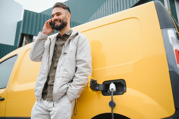 Bebaarde blanke man die naast een elektrische auto staat die wordt opgeladen en tijdsaanpassingen maakt op een smartphone