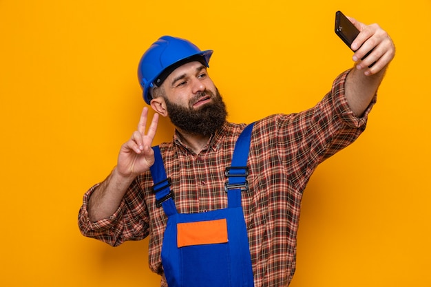Bebaarde bouwer man in bouw uniform en veiligheidshelm doen selfie met smartphone glimlachend vrolijk tonen v-teken staande over oranje achtergrond