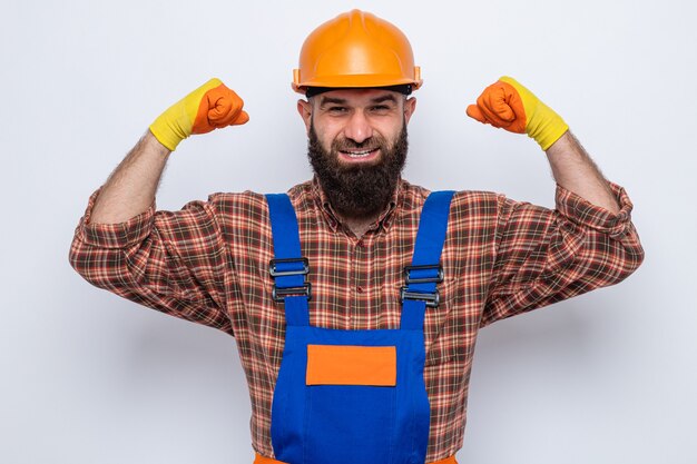 Bebaarde bouwer man in bouw uniform en veiligheidshelm dragen van rubberen handschoenen kijken camera glimlachend vertrouwen verhogen vuisten als een winnaar permanent op witte achtergrond