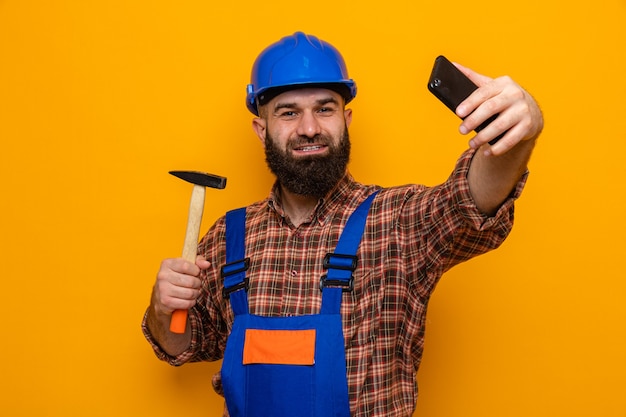 Bebaarde bouwman in bouwuniform en veiligheidshelm met hamer die selfie maakt met een vrolijk lachende smartphone