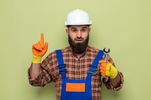 Bebaarde bouwman in bouwuniform en veiligheidshelm met rubberen handschoenen die een moersleutel vasthoudt en er verbaasd uitziet terwijl hij met de wijsvinger naar de zijkant wijst