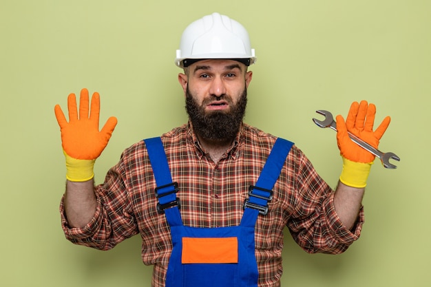 Bebaarde bouwman in bouwuniform en veiligheidshelm met rubberen handschoenen die een sleutel vasthoudt en er verward uitziet met opgeheven armen raising