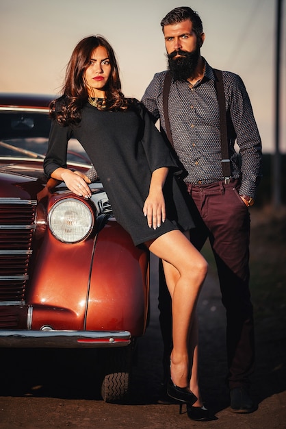 Bebaarde brunette man in een shirt en broek met bretels knuffelt een slank jong meisje met lang haar in een zwarte korte jurk in de buurt van een bruine retro auto op een donkere veldachtergrond