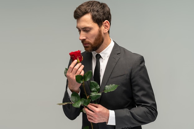 Bebaarde brunette man in formeel pak staande houden rood roze bloem ruiken en genieten met gesloten ogen Indoor studio shot geïsoleerd op grijze achtergrond