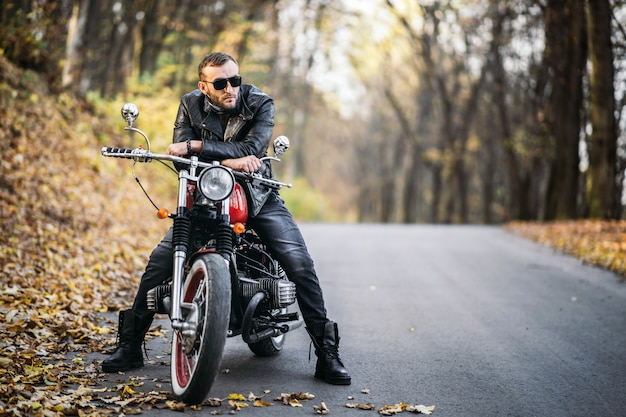 Bebaarde brute man in zonnebril en lederen jas zittend op een motorfiets op de weg in het bos