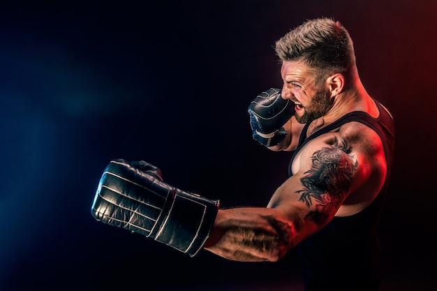 Bebaarde getatoeëerde sportman muay thai bokser in zwart onderhemd en bokshandschoenen vechten op donkere achtergrond met rook. Sport concept.