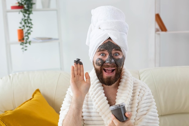 Bebaarde grappige man die plezier heeft met een cosmetisch masker op zijn gezicht gemaakt van zwarte klei. Mannen huidverzorging, humor en spa thuis concept