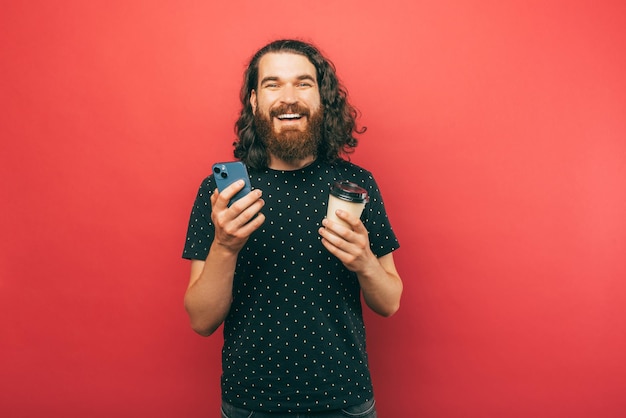 Bebaarde hipster houdt zijn koffie en zijn telefoon glimlachend naar de camera op rode achtergrond